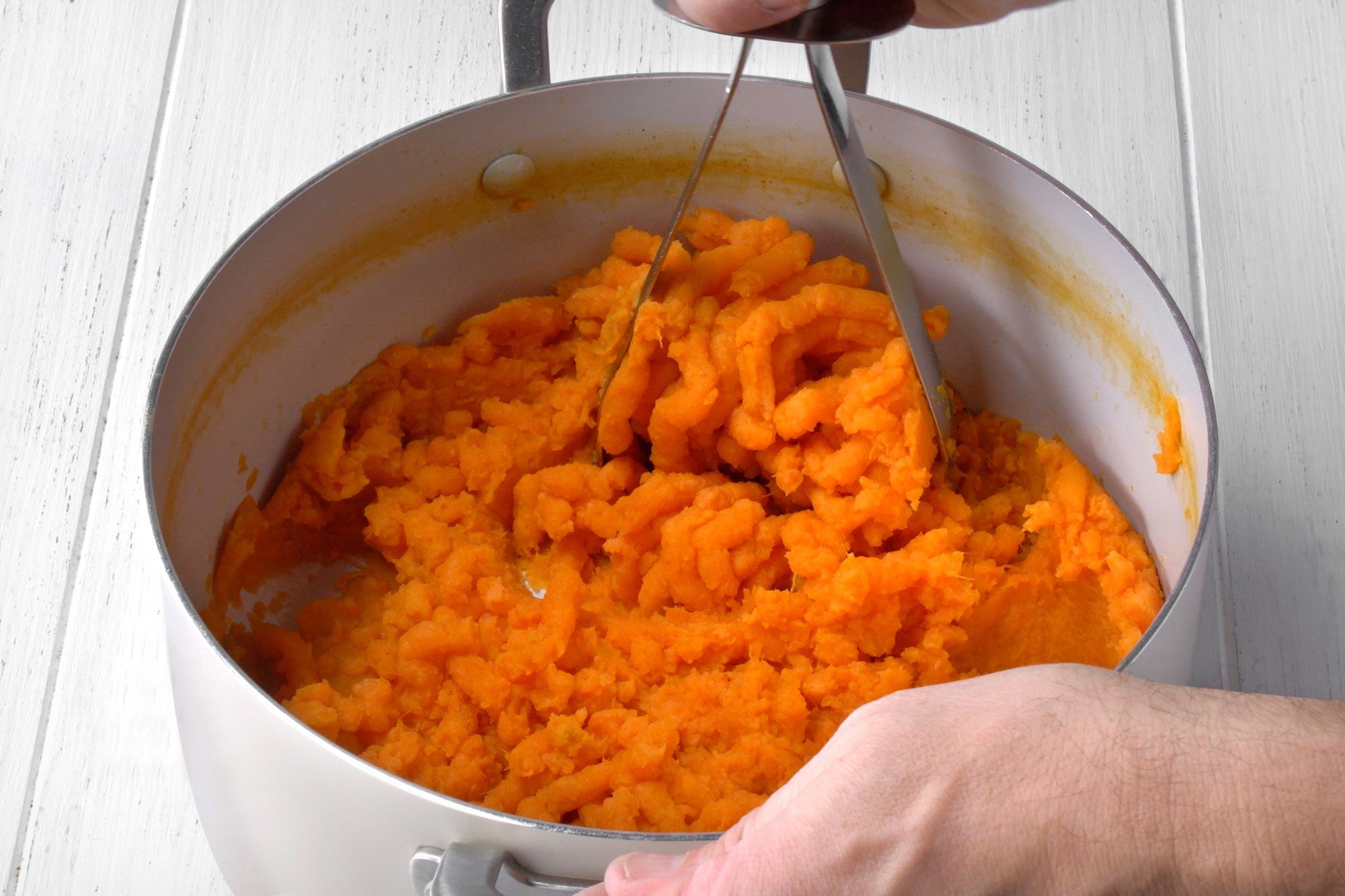 3/4th shot of a person mashing cooked sweet potatoes in a pot with a potato masher; the person is holding the potato masher with their right hand, and their left hand is resting on the side of the pot; the background is a white wooden surface