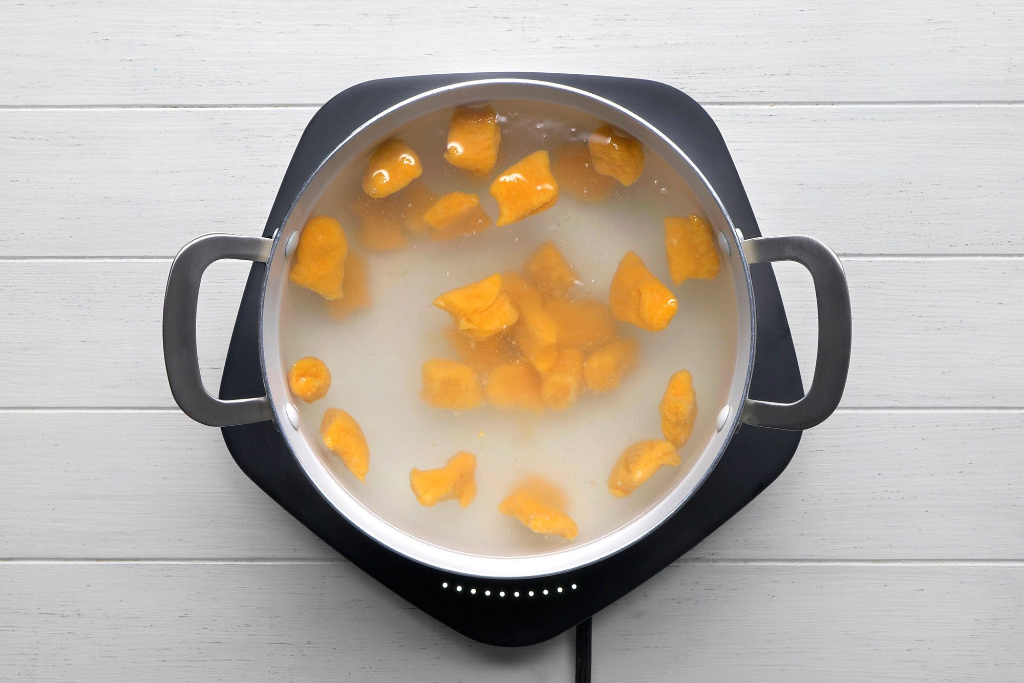 overhead shot of a pot of sweet potato gnocchi simmering on a stovetop; the pot is sitting on a black burner with white dots; the gnocchi are floating in the water and are a bright orange color; the pot is on a white wooden surface