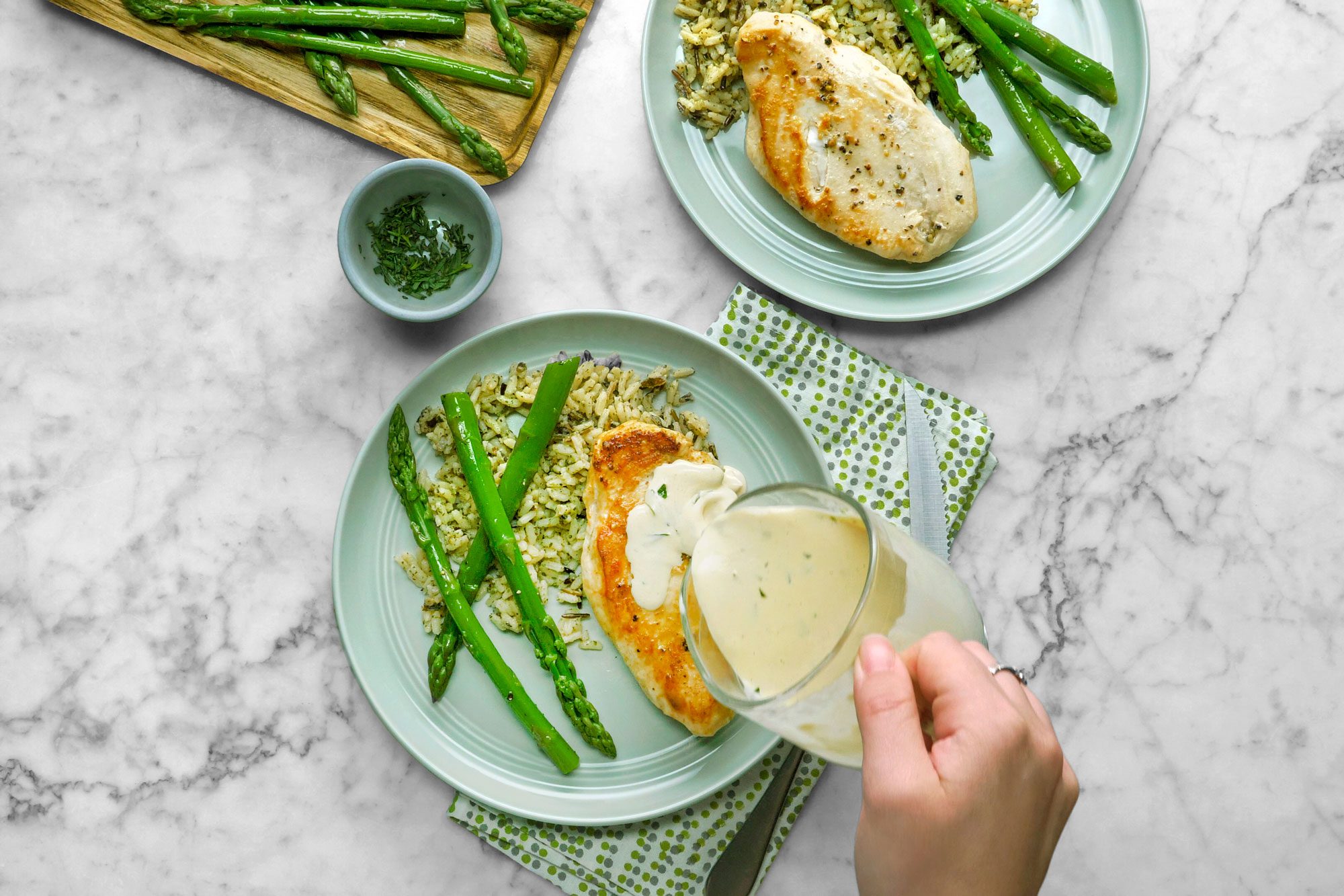 Top view shot of Chicken with Tarragon Sauce; serve on each plates with chicken; sprinkle with remaining tarragon; asparagus; napkin; cutlery; marble surface
