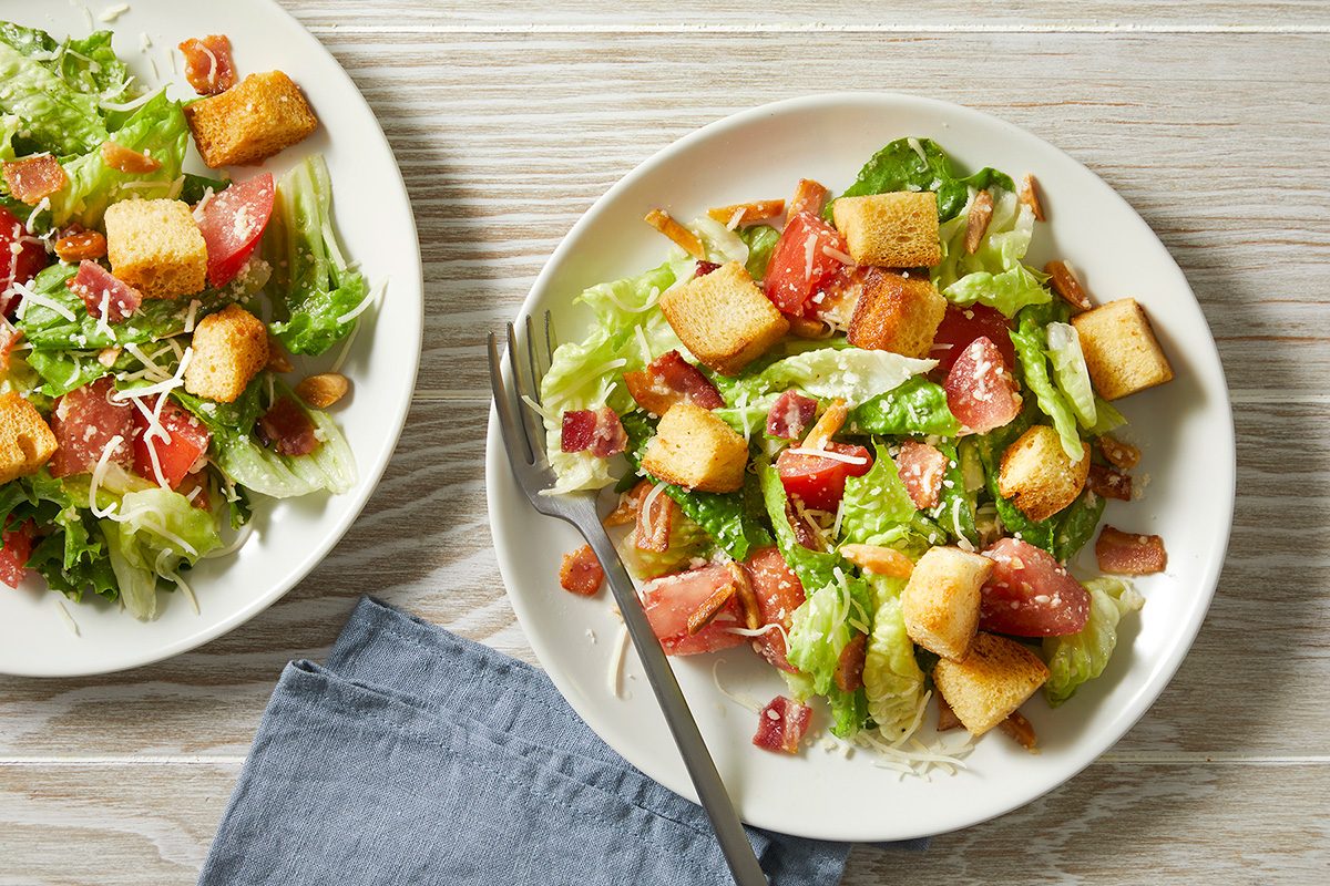 Two plates of salad on a wooden table, featuring lettuce, tomatoes, croutons, and bacon bits. A fork rests on one plate, with a blue napkin beside it.