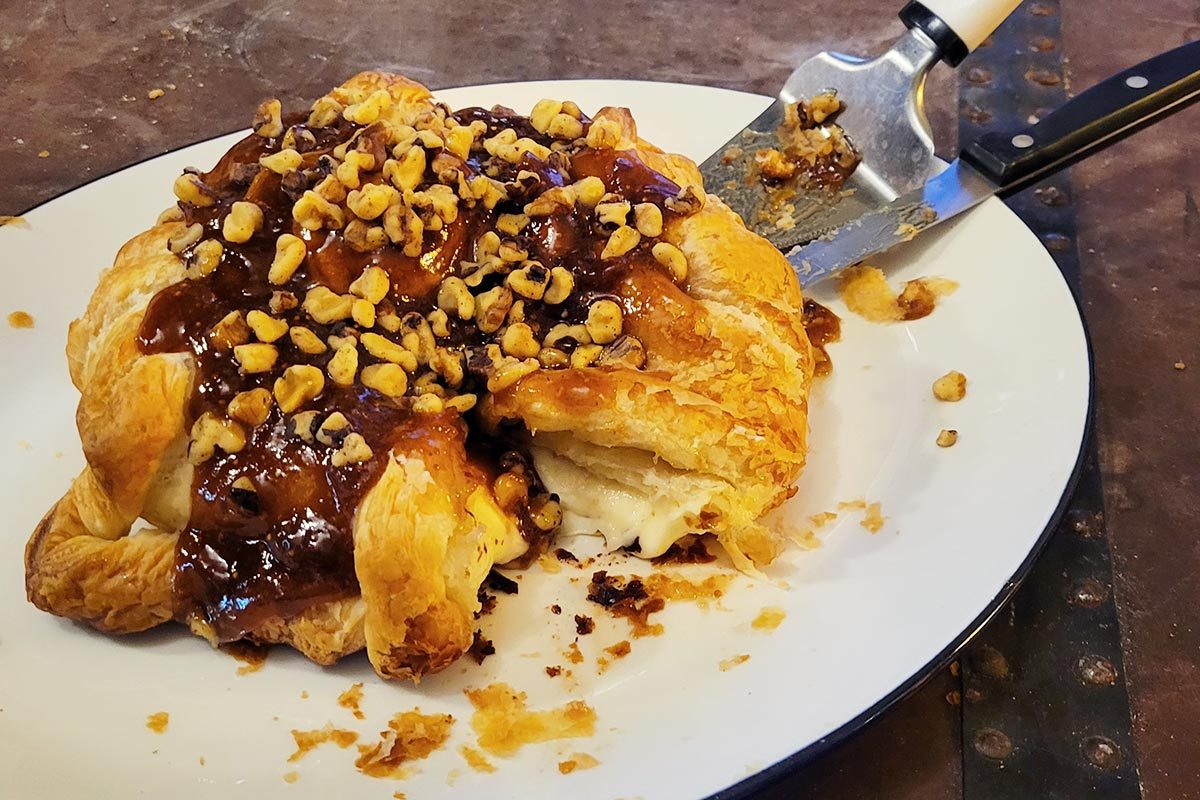 A baked wheel of brie cheese in a puff pastry, topped with jam and nuts with a knife and a serving spoon on a white plate 
