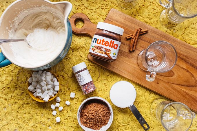 Bowl of whipped cream, bowl of marshmallows, bowl of cocoa powder, measuring cup of sugar, container of sprinkles, cinnamon sticks, jar of nutella, and several empty glasses sit on a yellow lace tablecloth and wooden cutting board