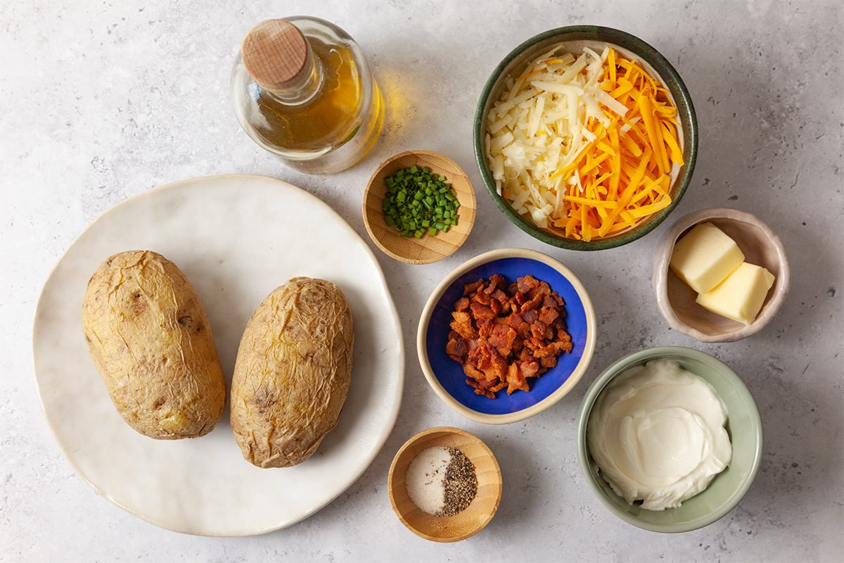 Ingredients for loaded baked potatoes arranged on a surface: two whole baked potatoes, grated cheese, chopped chives, crispy bacon bits, butter, sour cream, salt, pepper, and a bottle of oil.