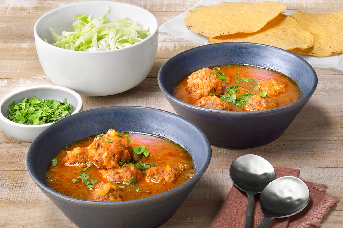 Two bowls of meatball soup garnished with cilantro are on a wooden table. Nearby, there's a small bowl of chopped cilantro, a larger bowl of shredded lettuce, two spoons, a cloth napkin, and some flat, crispy tortillas.