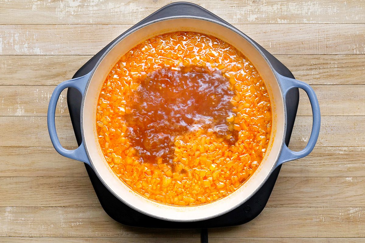 A pot on a stovetop contains a simmering orange liquid mixture with chunks, possibly a soup or sauce, in a light-colored saucepan. The pot has two handles and sits on a wooden surface.