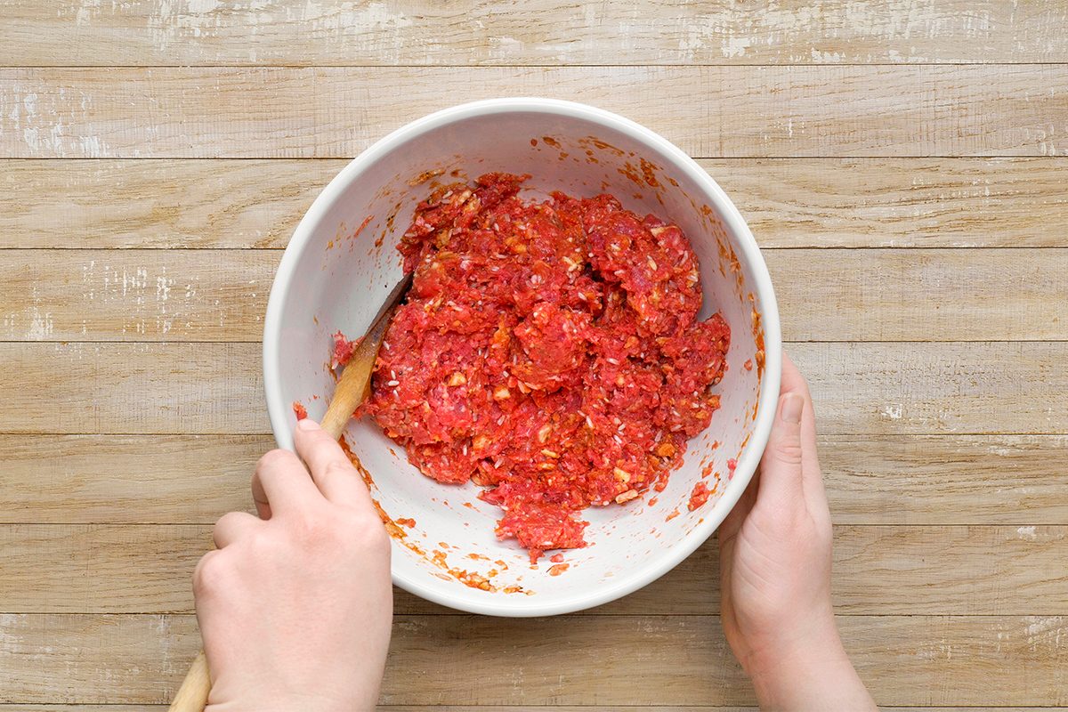 Two hands are mixing a red ground meat mixture with a wooden spoon in a white bowl. The bowl is on a wooden surface.