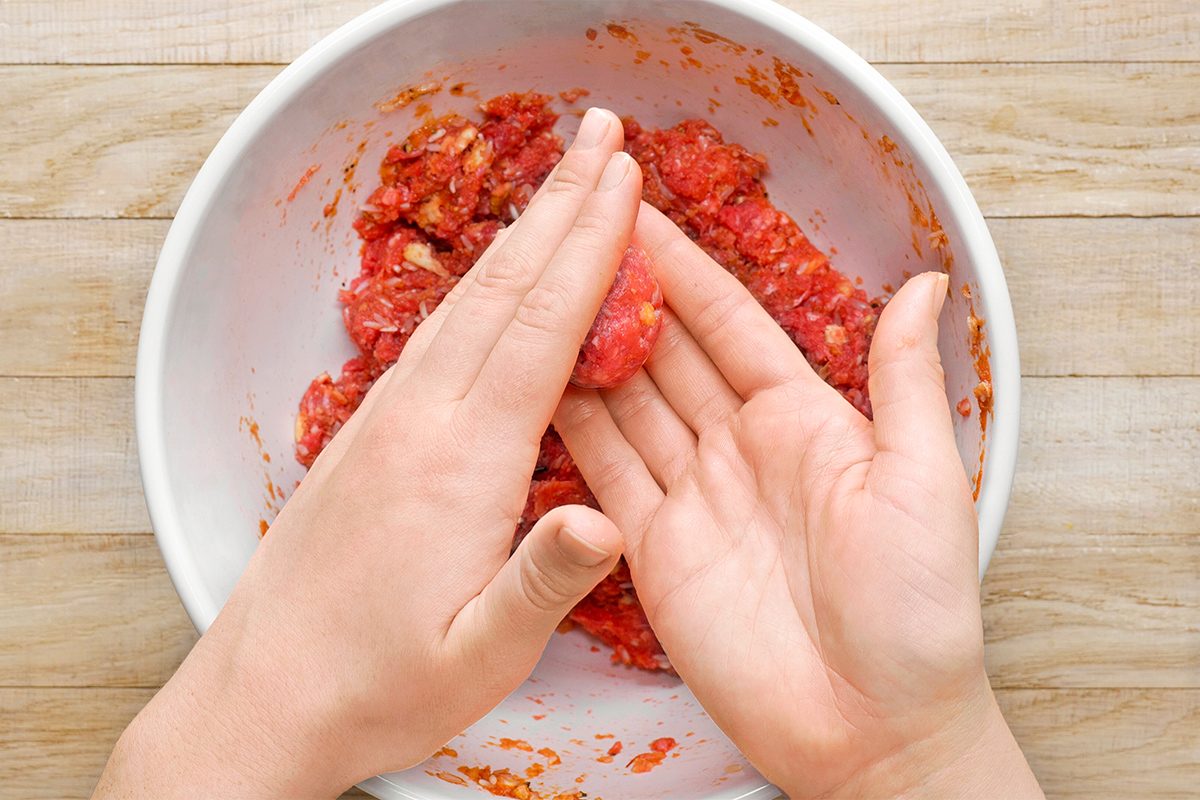 Hands shaping ground meat mixture into a ball over a white bowl on a wooden surface.