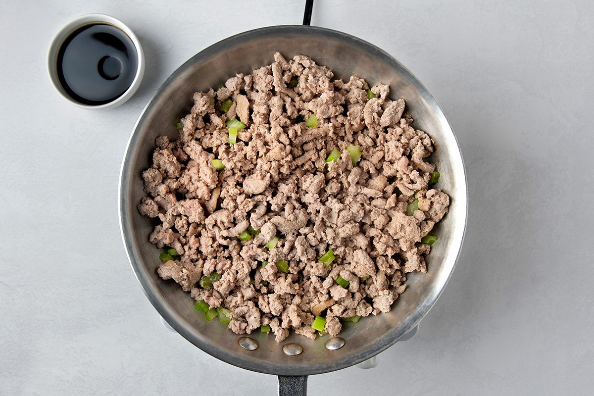 A pan filled with cooked ground turkey mixed with green pepper pieces. A small bowl containing a dark sauce is placed on the side, on a light gray surface.