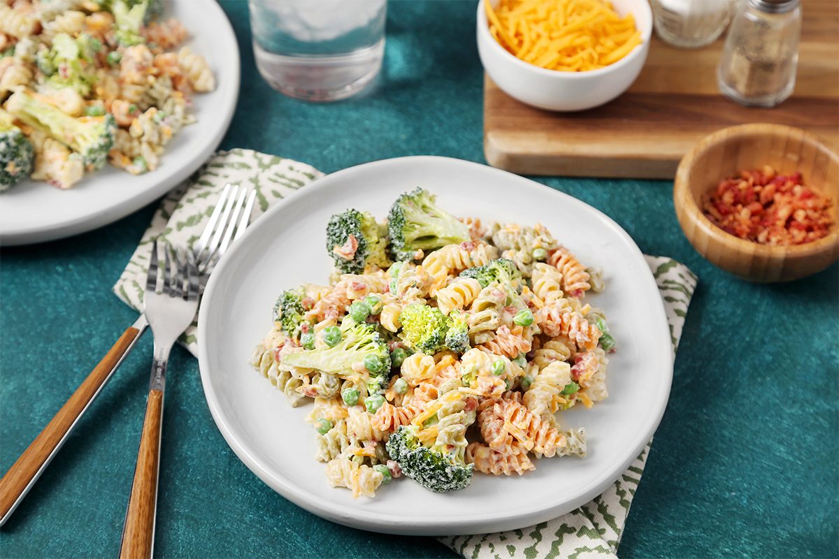 A plate of rotini pasta salad with broccoli, carrots, and dressing is on a table. Nearby are bowls of shredded cheese and bacon bits, a glass of water, and utensils. The table surface is green, and a patterned cloth napkin is under the plate.
