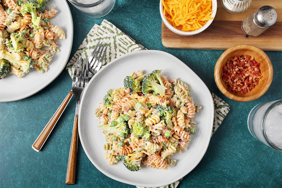 A plate of creamy pasta salad with rotini, broccoli, and dressing is on a table. Nearby are bowls of shredded cheese and bacon bits, along with a salt and pepper shaker. Forks and a patterned napkin are also visible.