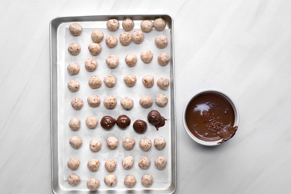 A baking tray with rows of small dough balls on parchment paper. Some balls are coated in chocolate. A bowl of melted chocolate is next to the tray on a white surface.