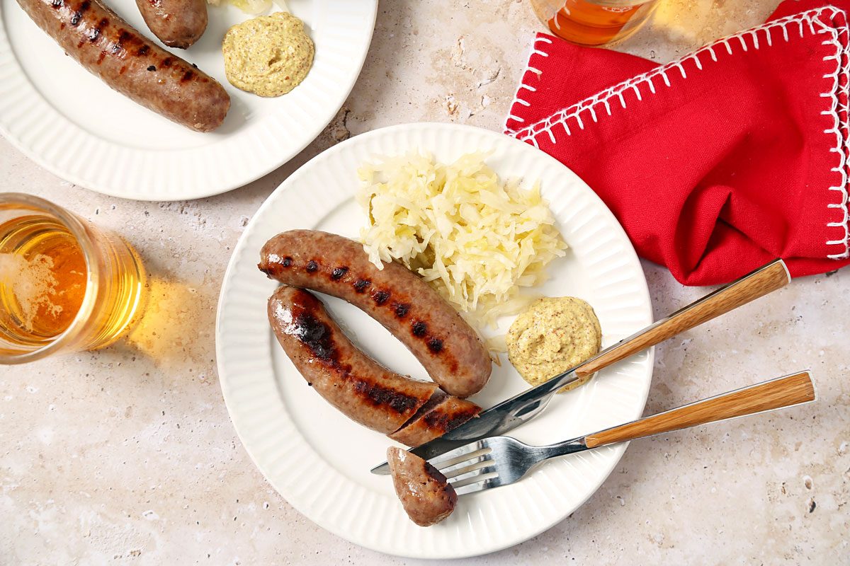 overhead shot of a delicious meal of grilled sausages, sauerkraut, and mustard; two white plates are on a light brown table; one plate has two grilled sausages and a dollop of mustard; the other plate has two grilled sausages, a serving of sauerkraut, and a dollop of mustard; a fork and knife lay on the plate with the sauerkraut; a glass of beer is on the table; a red napkin with white trim is on the table
