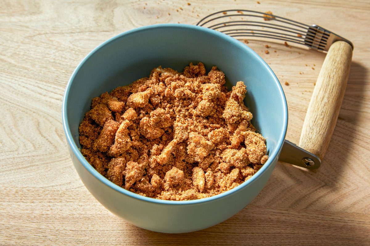 3/4th shot of a bowl filled with a mixture of crushed biscuits, sugar, and cinnamon; the bowl is light blue and rests on a wooden surface; a pastry blender is on the right side of the bowl