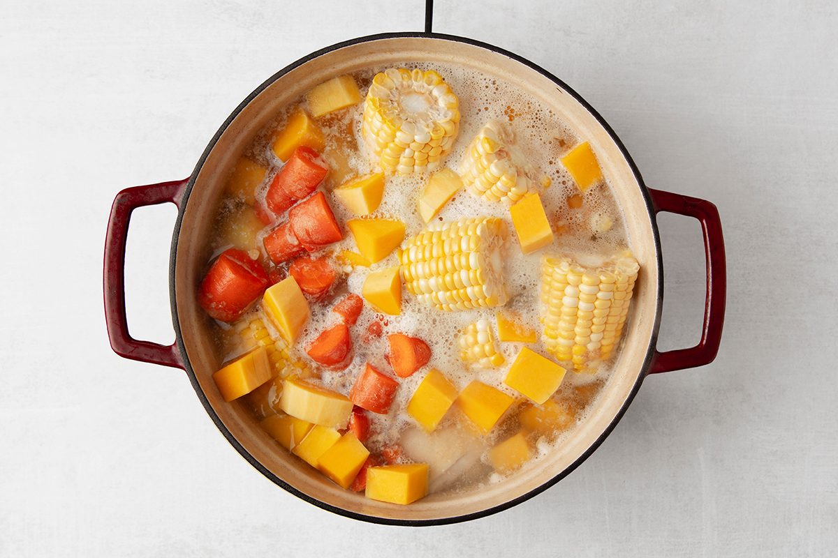 A pot filled with bubbling soup, containing pieces of corn on the cob, chunks of squash, and slices of red bell pepper, on a white countertop. The pot has two red handles.