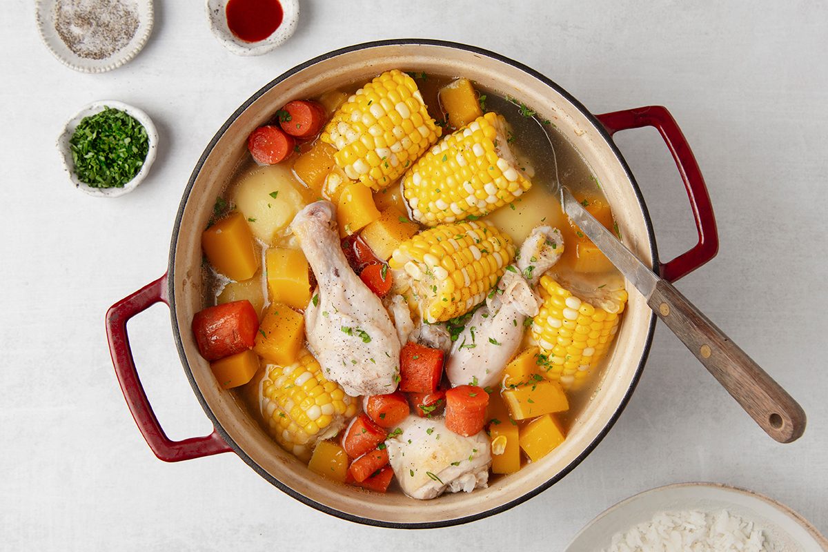 A pot of stew with chicken drumsticks, corn on the cob pieces, carrots, and squash. It is set on a light surface, accompanied by small bowls of herbs, spices, and a small plate of flour. A knife rests on the pot's rim.