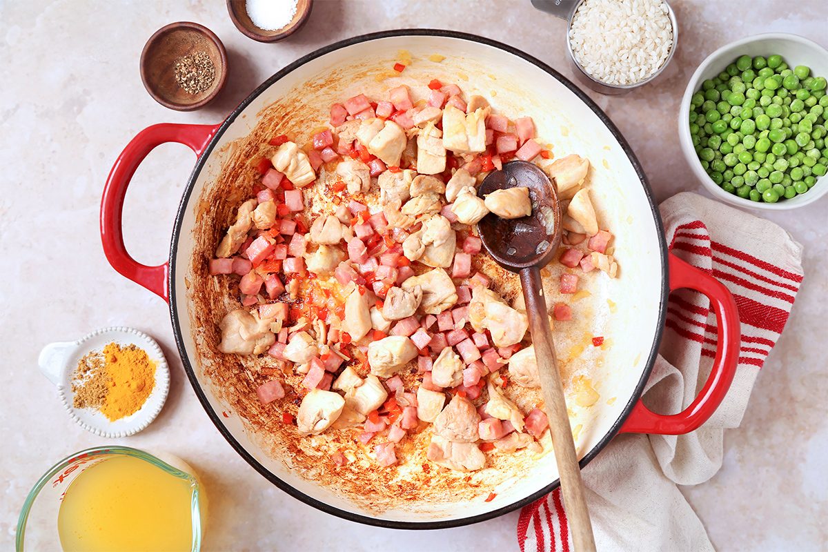 A red pot filled with diced chicken and ham, being stirred with a ladle. Surrounding the pot are bowls of peas, rice, and spices, as well as a glass of juice. A red-striped towel is placed beside the pot.