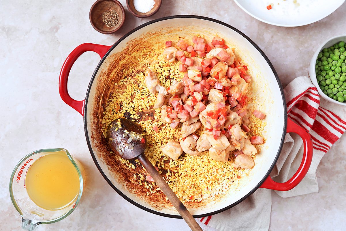 A red Dutch oven on a light countertop contains cooked chicken pieces and rice, with diced tomatoes on top. A wooden spoon rests inside. Nearby, there are bowls of salt, pepper, green peas, and a measuring cup with broth.