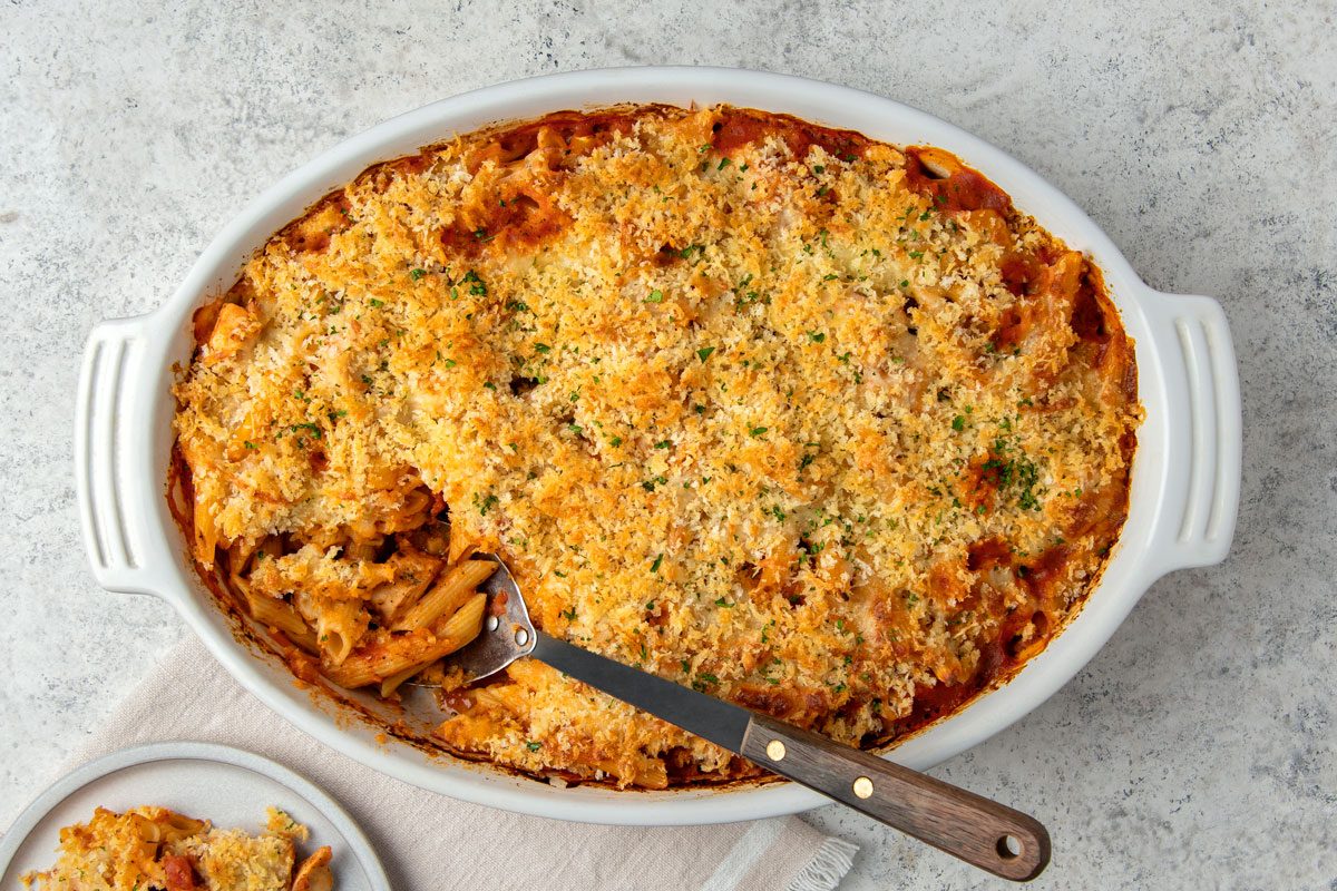 Overhead shot of Chicken Parmesan Casserole; in a baking pan; serving spoon; served on a small plate; napkin; marble surface;