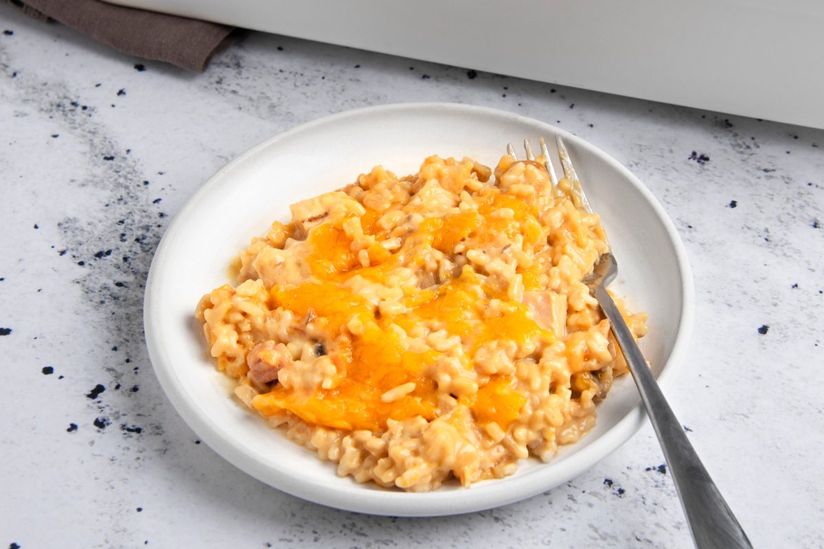 3/4th shot of a white plate with a serving of cheesy rice casserole on it; a fork is laid beside the plate; the casserole looks creamy and cheesy, with rice and some meat mixed in; the plate is set on a white surface with black specks
