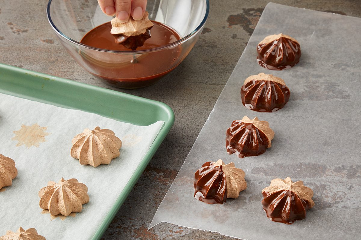 A hand dips meringue cookies into a bowl of melted chocolate. Some meringues are set on wax paper with chocolate coating partially covering them, while others are on a green tray lined with parchment paper.