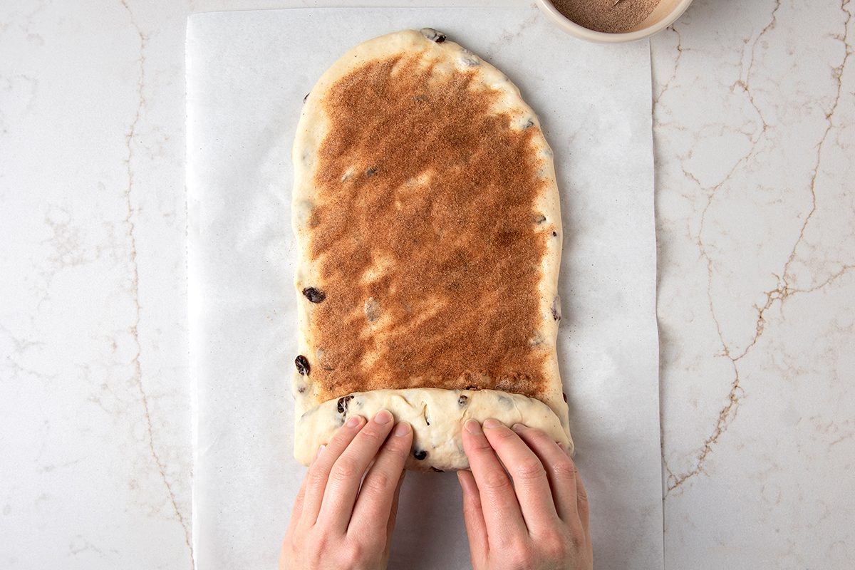Hands rolling cinnamon raisin dough on parchment paper, with a small bowl nearby on a marble surface.