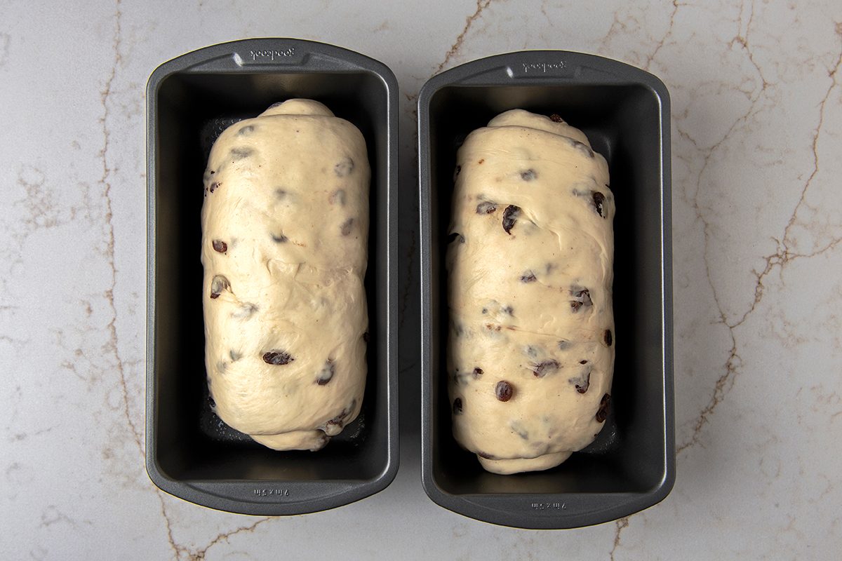 Two loaf pans sit side by side on a marble surface, each containing uncooked dough mixed with raisins. The dough is risen and ready to be baked, with a light, smooth texture visible on top.