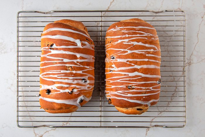 Two loaves of golden-brown bread topped with white glaze sit on a cooling rack. The bread is dotted with dark pieces, possibly raisins, and the rack is placed on a marble surface.