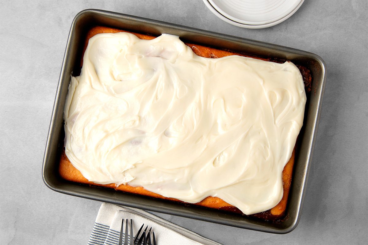 A rectangular cake in a metal baking pan covered with smooth, white frosting. It rests on a light gray surface next to a folded napkin with forks and a white plate with a paper towel.