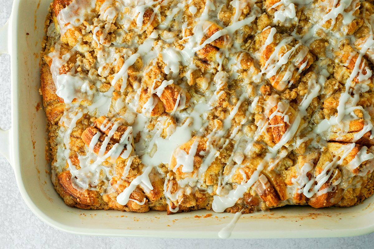 A close-up of a baked apple cinnamon bread pudding in a rectangular dish. The dessert is topped with a drizzle of white icing, showcasing a golden-brown crust with visible apple chunks and pieces of bread.