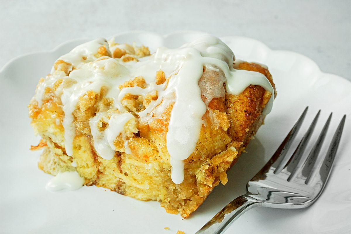 A close-up of a piece of cinnamon roll casserole on a white plate, drizzled with white icing. A fork is placed beside the dessert, ready for use. The background is a soft, out-of-focus white surface.