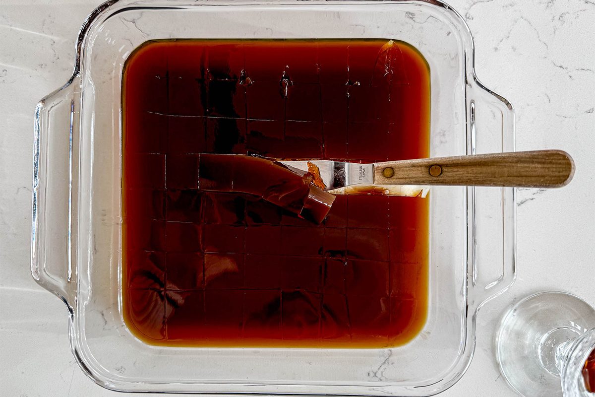 A square glass dish filled with brown gelatin-like coffee jelly, with a knife placed on top of it. The dish is on a white marble countertop, and a small part of a glass is visible on the right side.