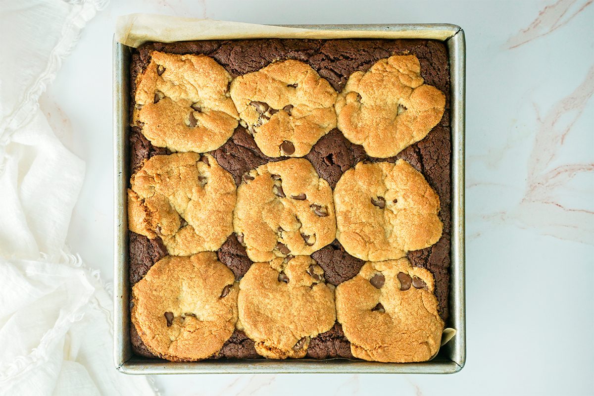 A square baking pan filled with freshly baked brookies, a combination of brownies and cookies. The top is golden brown with chocolate chip cookie dough spread unevenly across the brownie base. The pan is on a light marble surface.