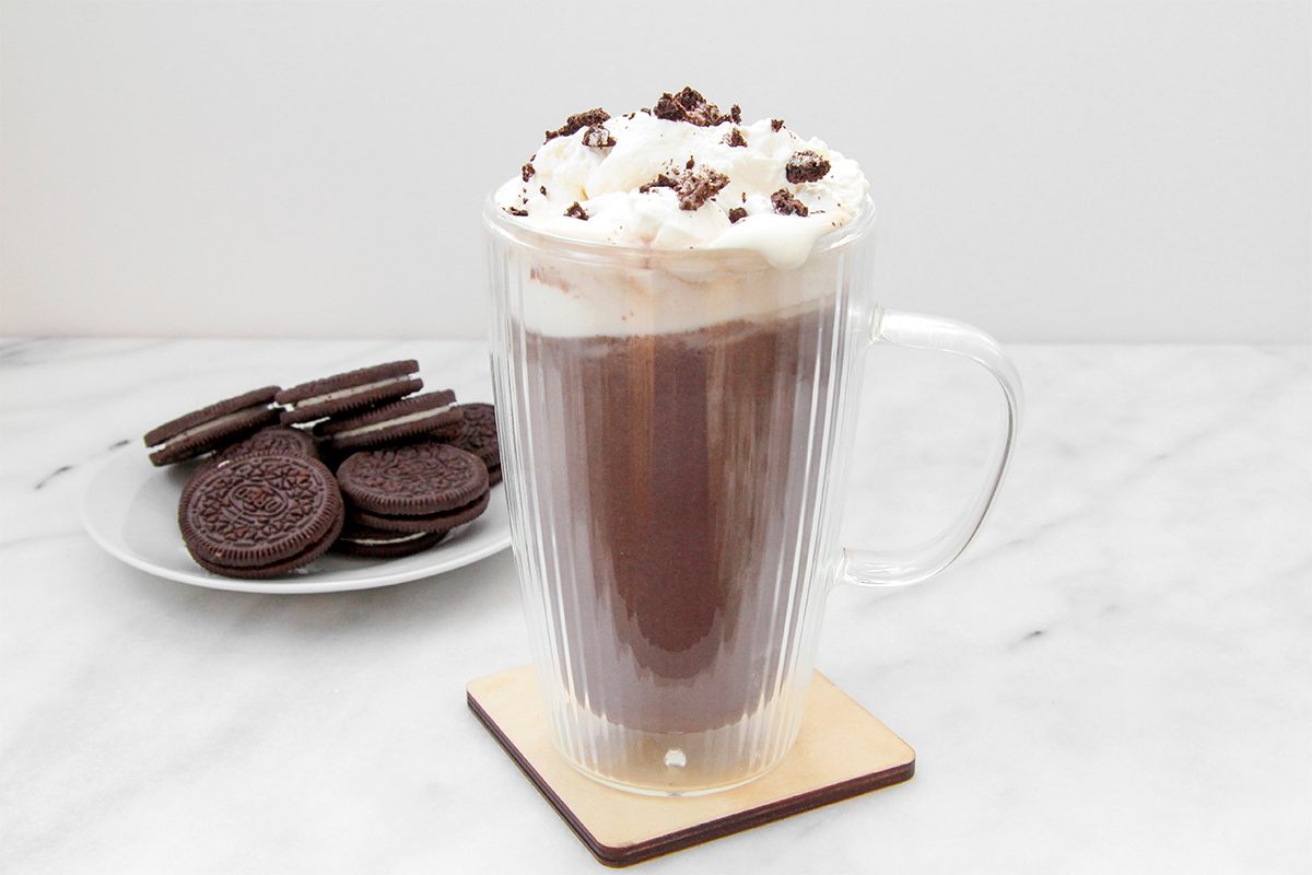 A glass mug of hot chocolate topped with whipped cream and chocolate shavings sits on a wooden coaster. In the background, there's a plate stacked with chocolate sandwich cookies on a marble surface.