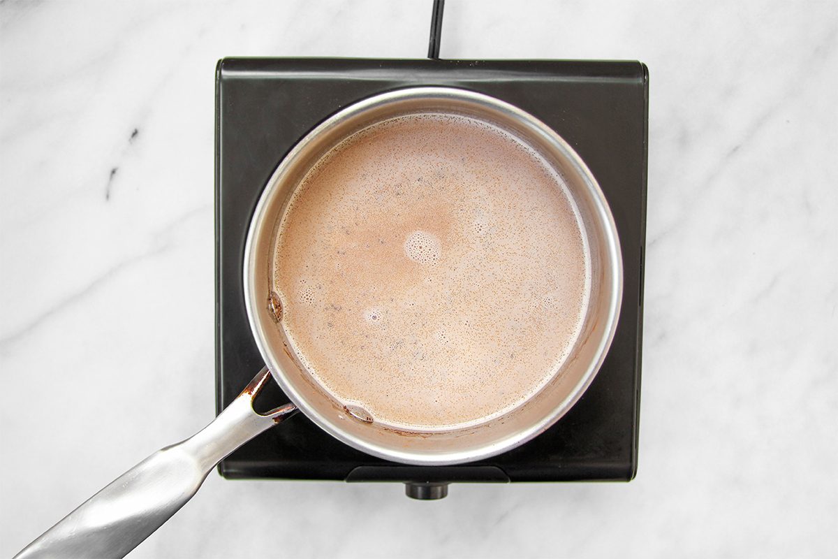 A pot of hot chocolate is being heated on a black stove top. The liquid is a light brown color, and the stovetop is placed on a white marble surface.
