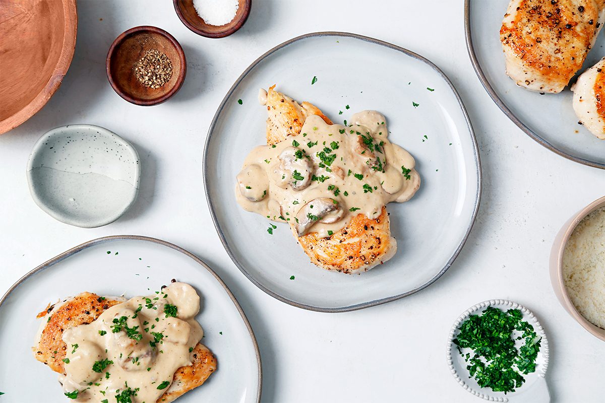 Plates of chicken topped with creamy mushroom sauce and garnished with chopped herbs are arranged on a white table. Accompanied by small bowls of salt, pepper, chopped herbs, and sauce, along with a wooden bowl in the corner.