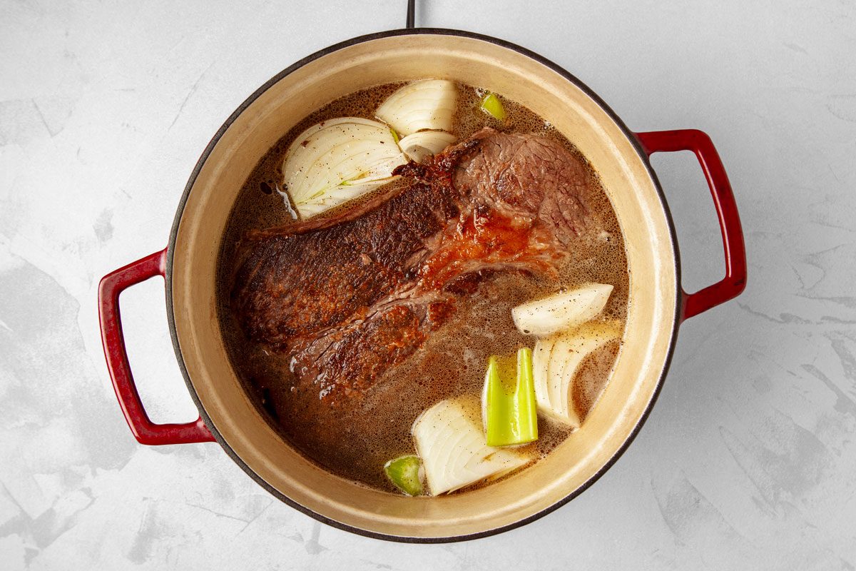 overhead shot of a Dutch oven pot roast; the pot is red and has a beige interior; the roast is a large piece of meat that has been browned and is sitting in a brown liquid with onions and celery; the onions are sliced and the celery is cut into large pieces