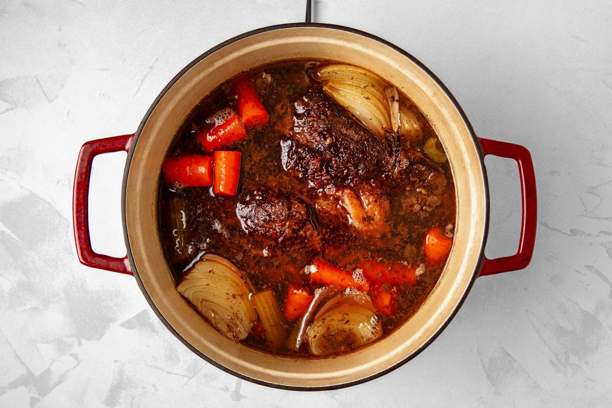 overhead shot of a Dutch oven pot roast cooking on the stovetop; a Dutch oven pot roast is a slow-cooked dish made with beef, vegetables, and broth; the pot is filled with a brown liquid that is likely broth or gravy, and there are chunks of meat, carrots, and onions in the liquid