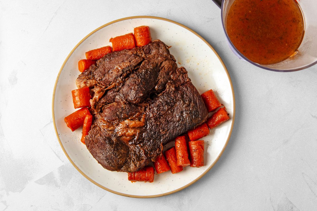 overhead shot of a Dutch oven pot roast; it consists of a large piece of browned pot roast with carrots around it; the roast and carrots are on a white plate with a brown rim; the plate is sitting on a white counter with a clear glass jar filled with brown gravy in the upper right-hand corner of the image