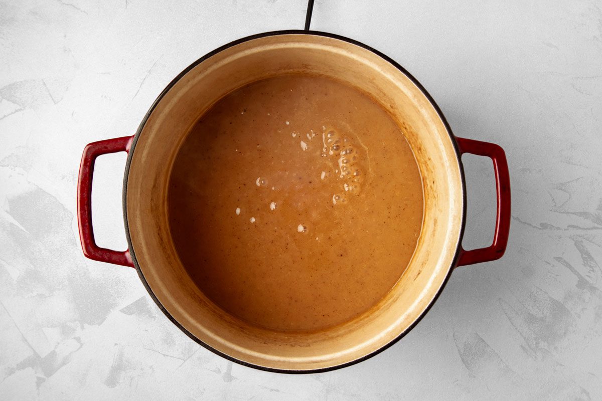 overhead shot of a red enamel cast iron pot filled with a brown liquid, likely a sauce or gravy; the pot is sitting on a white countertop with a textured surface