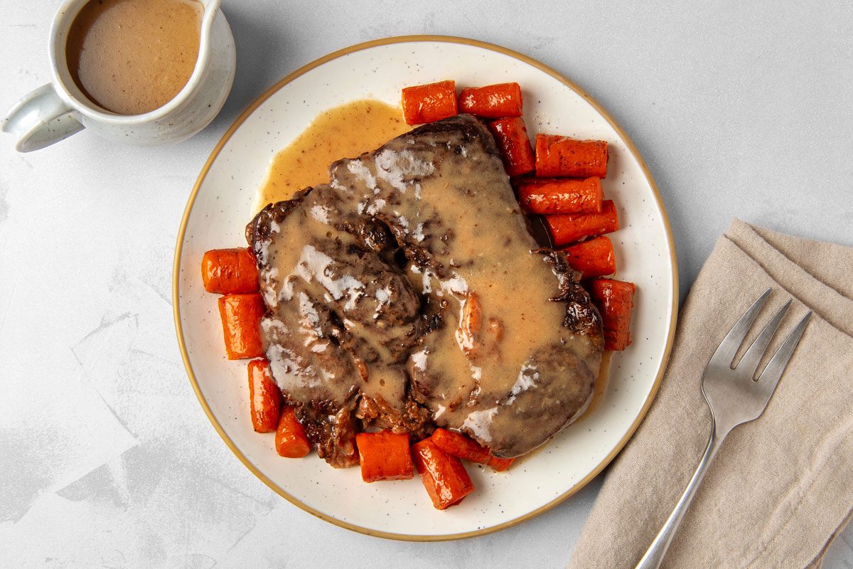 overhead shot of a plate of Dutch oven pot roast; the roast is a large cut of meat covered in a brown gravy; there is a small pitcher of gravy off to the side of the plate; a fork is placed on a tan napkin off to the side of the plate; the plate is on a white background