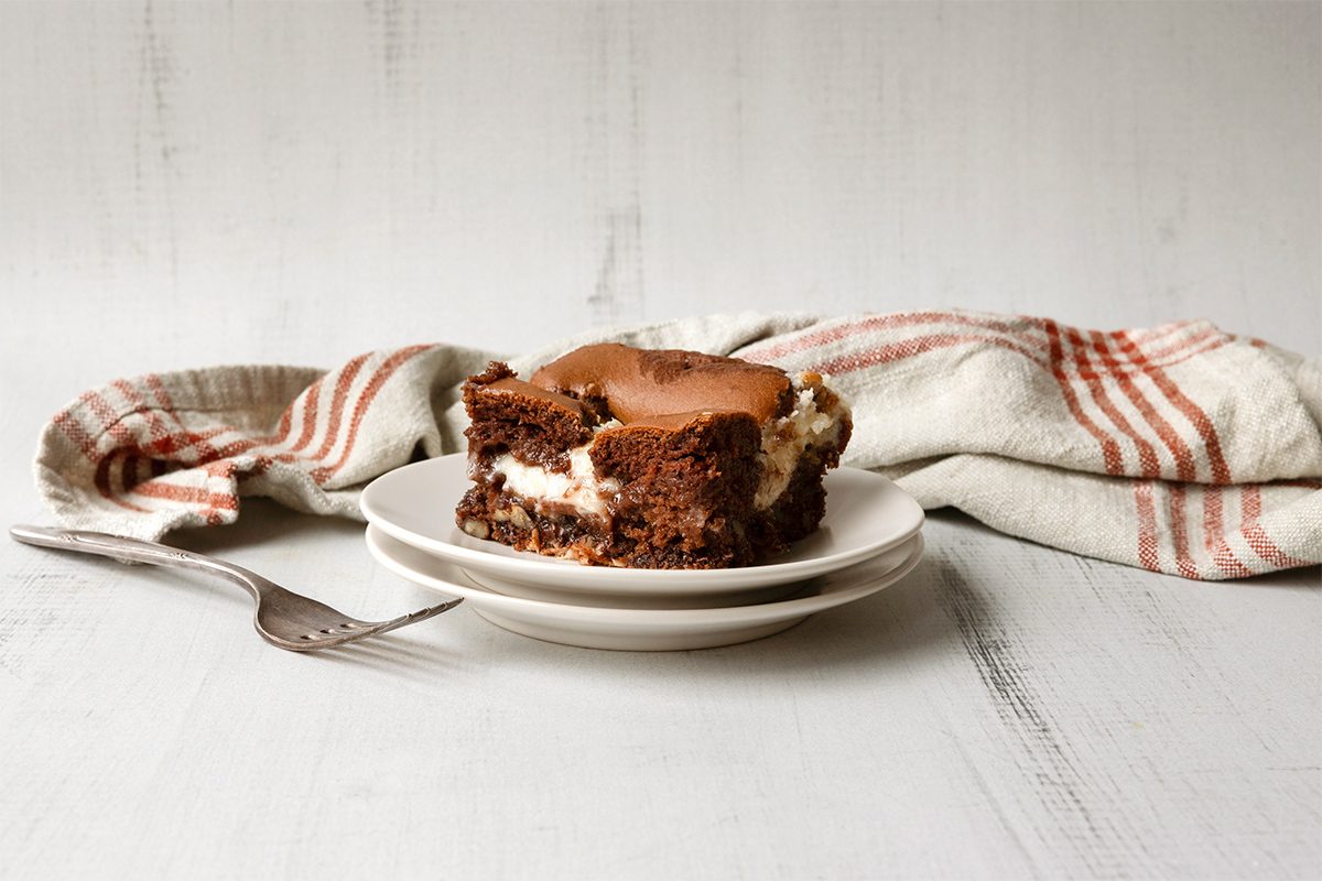 A slice of chocolate cake with cream filling on a white plate. A fork lies beside the plate on a white wooden surface. A striped kitchen towel is in the background.