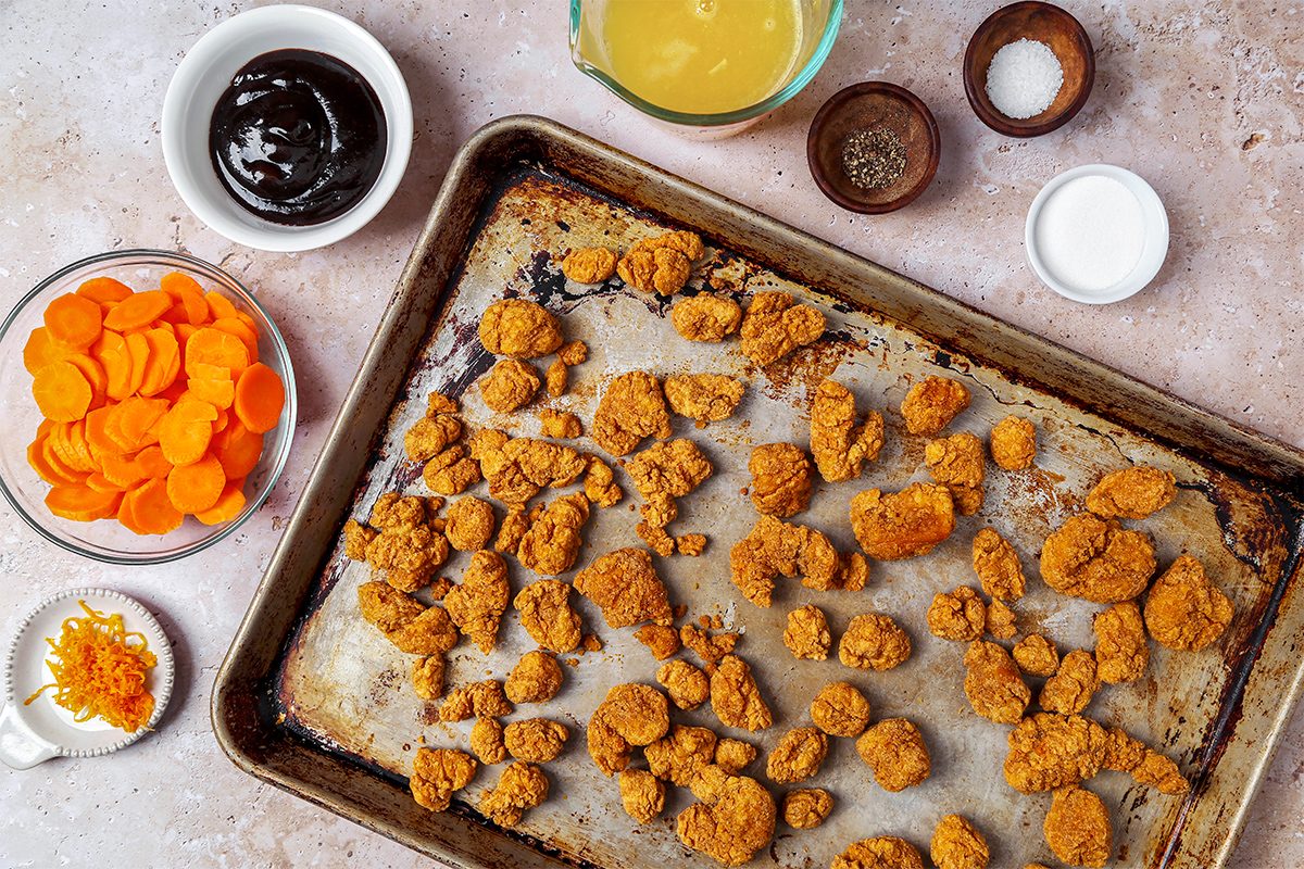 A baking sheet with crispy, golden-brown chicken nuggets surrounded by small bowls containing sliced carrots, soy sauce, a yellow sauce, seasonings, and citrus zest on a textured countertop.