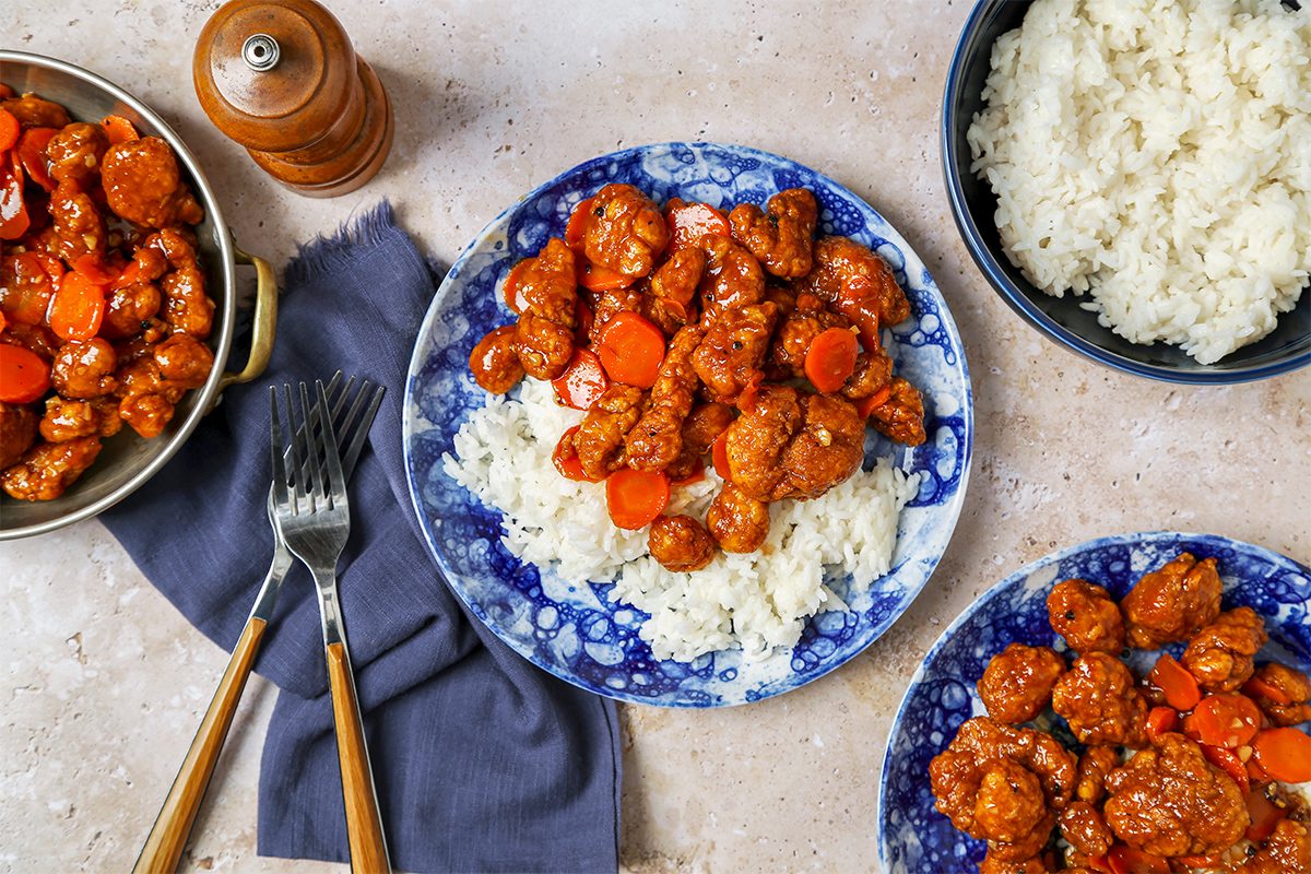 Plates of sweet and sour chicken with sliced carrots served on white rice. There are two plates on a blue-patterned dish, along with a bowl of rice, a serving dish of chicken, and cutlery resting on a blue napkin. A pepper grinder is in the background.