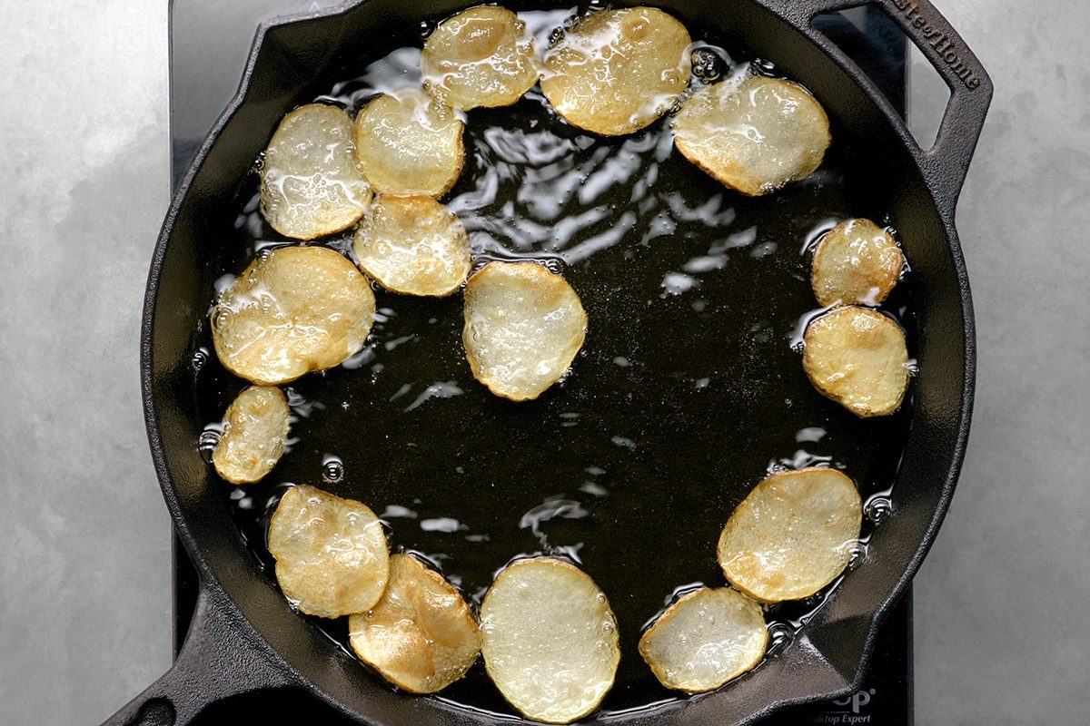 Potato slices frying in hot oil in a black cast iron skillet. The potato slices are golden brown and sizzling, surrounded by bubbles from the cooking oil. The skillet is on a gray stovetop surface.