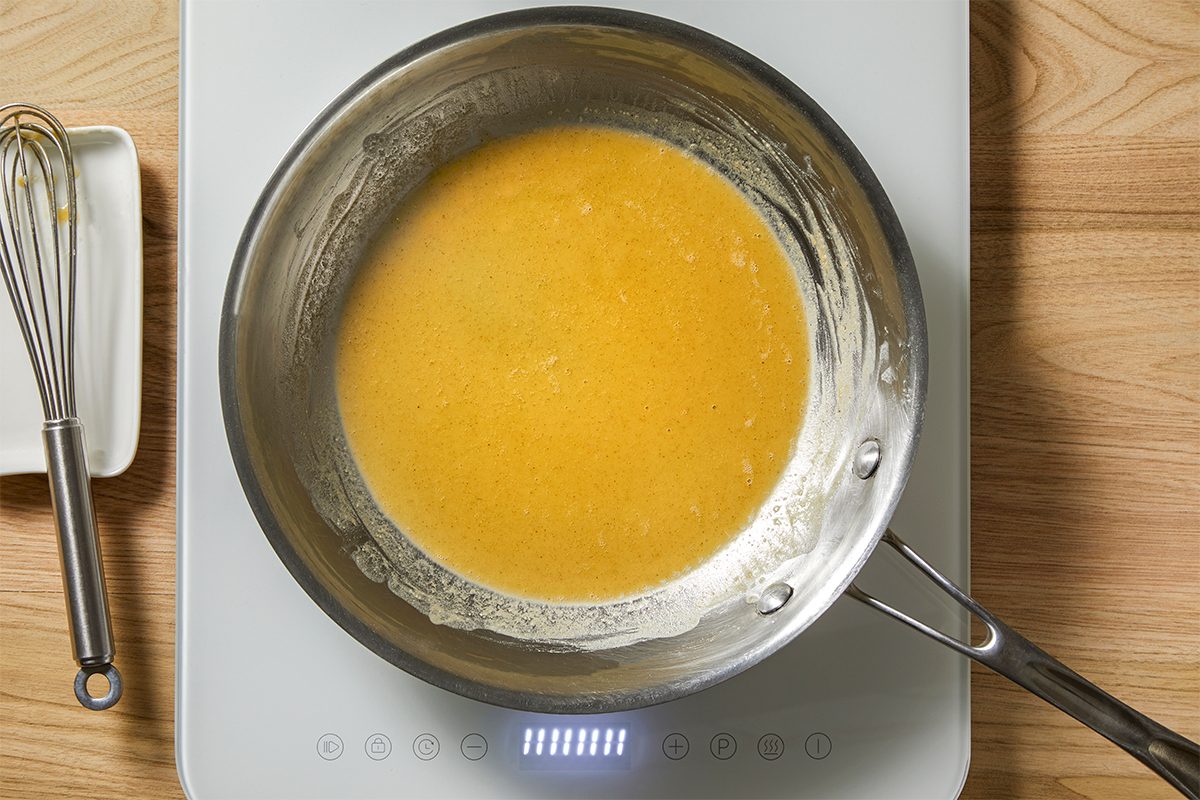A stainless steel pan on an induction stove contains a smooth, golden yellow sauce being cooked. A whisk rests on a white rectangular plate nearby, all set on a wooden surface.