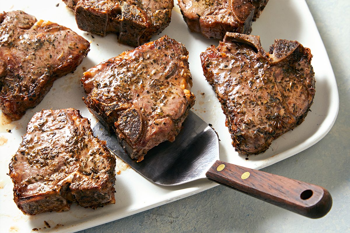 Overhead view of prepared lamb loin chops on a white serving tray and a serving spoon lifting one of them kept on a white marbletop.