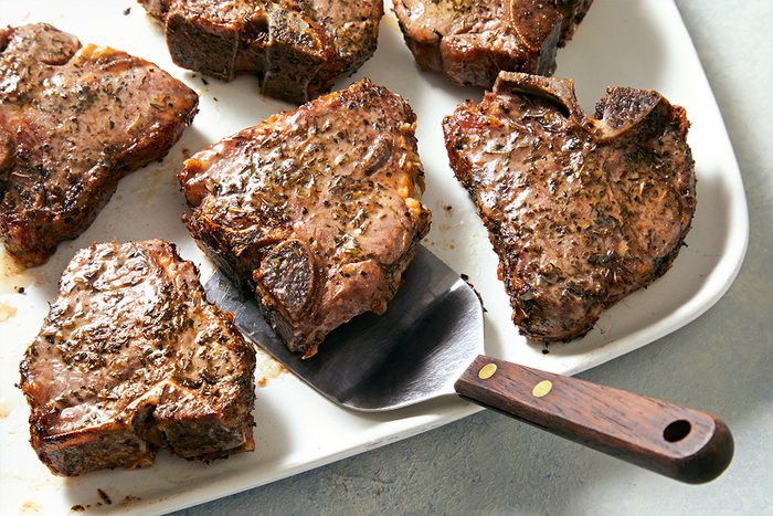 Overhead view of prepared lamb loin chops on a white serving tray and a serving spoon lifting one of them kept on a white marbletop.