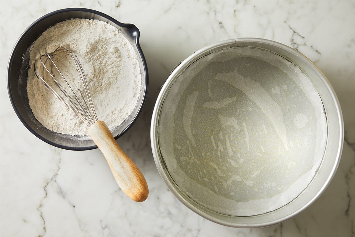 Overhead view of a bowl with a whisk containing dry mix on a white marble countertop. Next to it is a 9-inch springform pan lined with parchment paper.