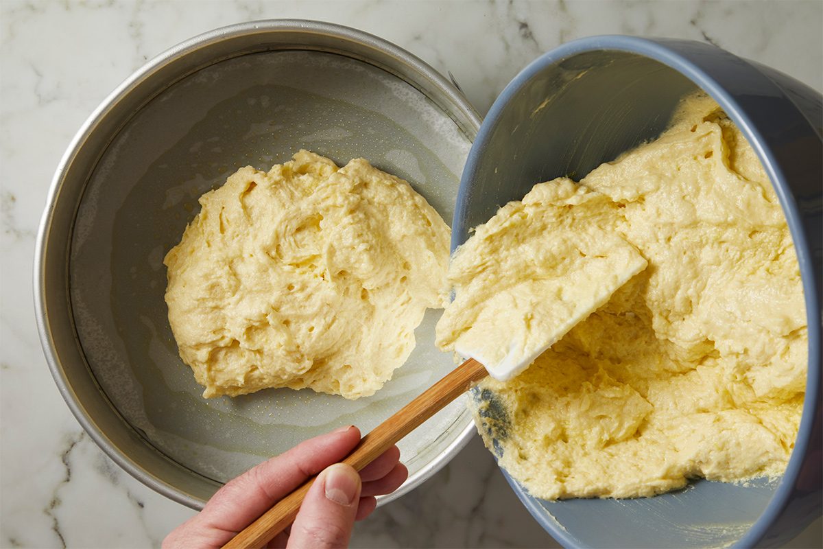 Overhead view of the mixed batter being poured into the lined springform pan with a spatula on a white marble countertop.