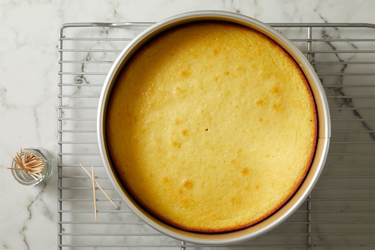 Overhead view of baked cake in the springform pan placed on a cooling rack on a white marble countertop with toothpicks in a container kept on the side.